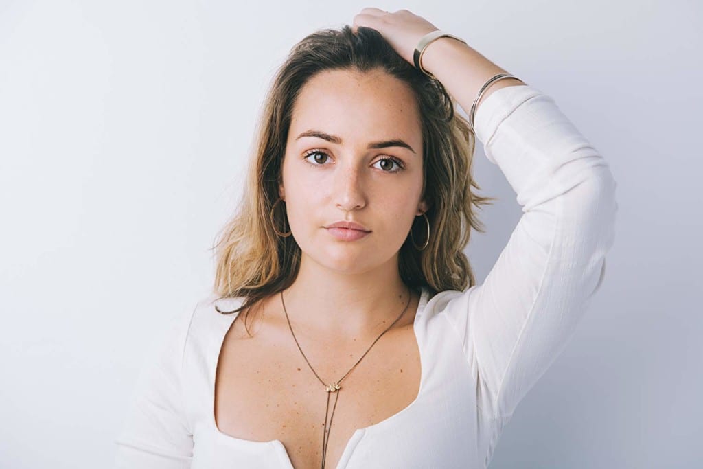 An actress looks directly into the camera. She has brown hair with highlights, hoop earrings, a necklack, and two bracelets. She is wearing a white long sleeved shirt and stands in front of a white background.