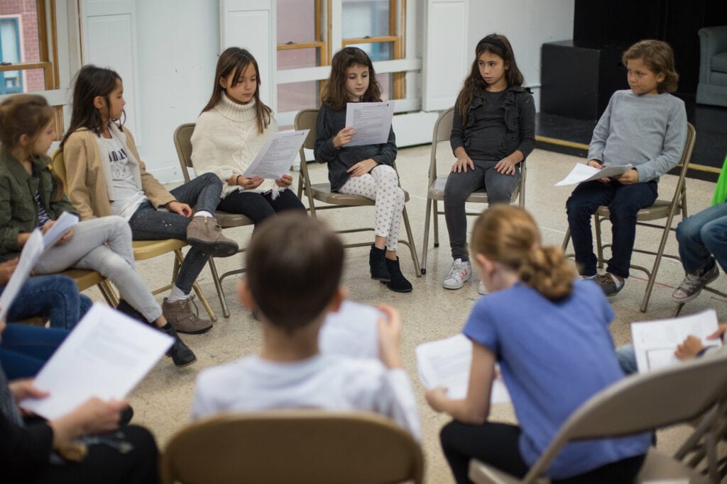 A group of young actors sit in a circle studying scripts