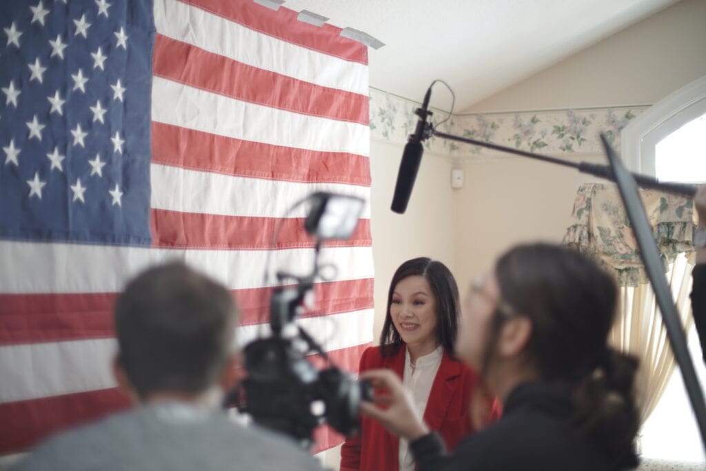 Leah Zhang with crew members around her and an American Flag on set behind.